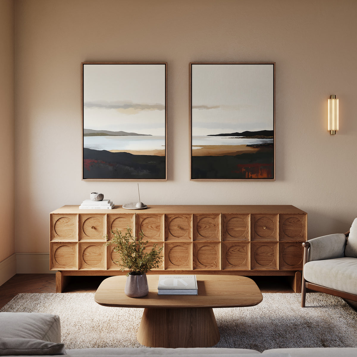 A modern living room features a wooden sideboard, the Nordic Waters - Set Of 2 prints above it, a coffee table with a book and vase, a light-colored armchair, and soft wall sconces for a calming decor.