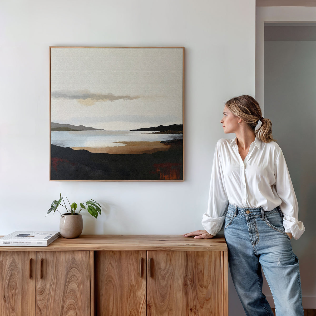 A woman in a white blouse and jeans leans against a wooden cabinet, gazing at the "Nordic Waters Canvas Art - Square," which depicts a serene lake and hills. A potted plant and books complete the sophisticated decor atop the cabinet.