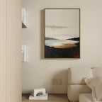 The Nordic Waters Canvas Art - Vertical features a coastal scene with dark hills and calm water in warm earth tones. Hanging on a beige wall, it complements a cream sofa and minimal shelf with books for a serene, stylish look.
