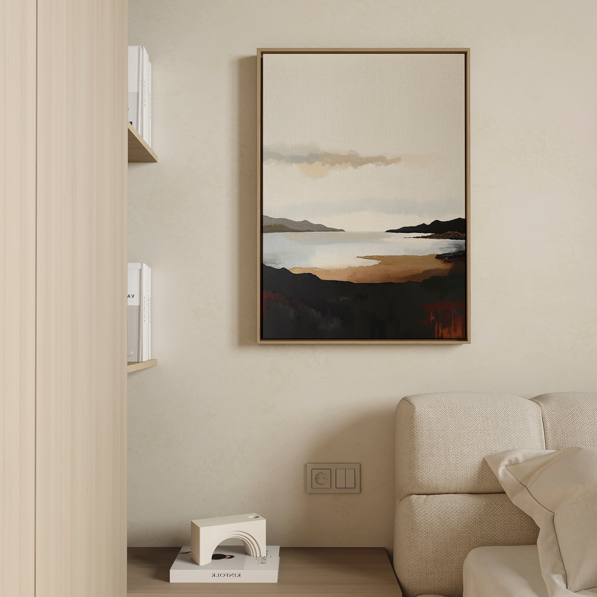 The Nordic Waters Canvas Art - Vertical features a coastal scene with dark hills and calm water in warm earth tones. Hanging on a beige wall, it complements a cream sofa and minimal shelf with books for a serene, stylish look.