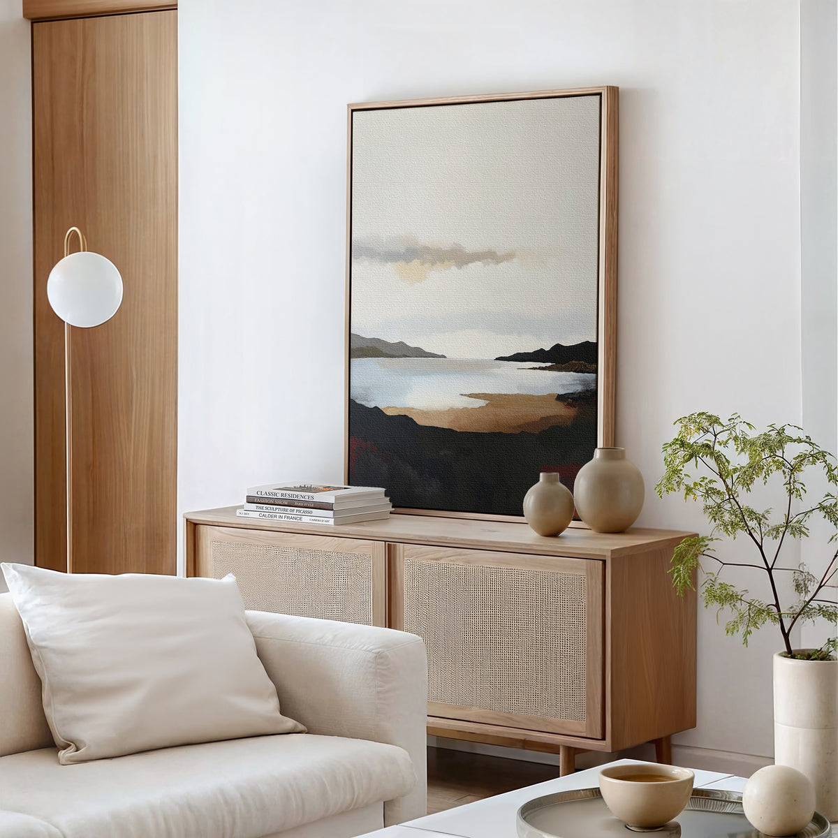 A minimal living room features a light wood sideboard with the Nordic Waters Canvas Art - Vertical, ceramic vases, a stack of books, a white armchair with pillow, a modern round floor lamp, and a small potted plant.