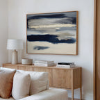 A modern living room features a light wood sideboard, white lamp, stacked books, neutral decor, and the Northern Tides Canvas Art - Horizontal—a striking abstract blue and beige painting—hanging above a cream sofa.