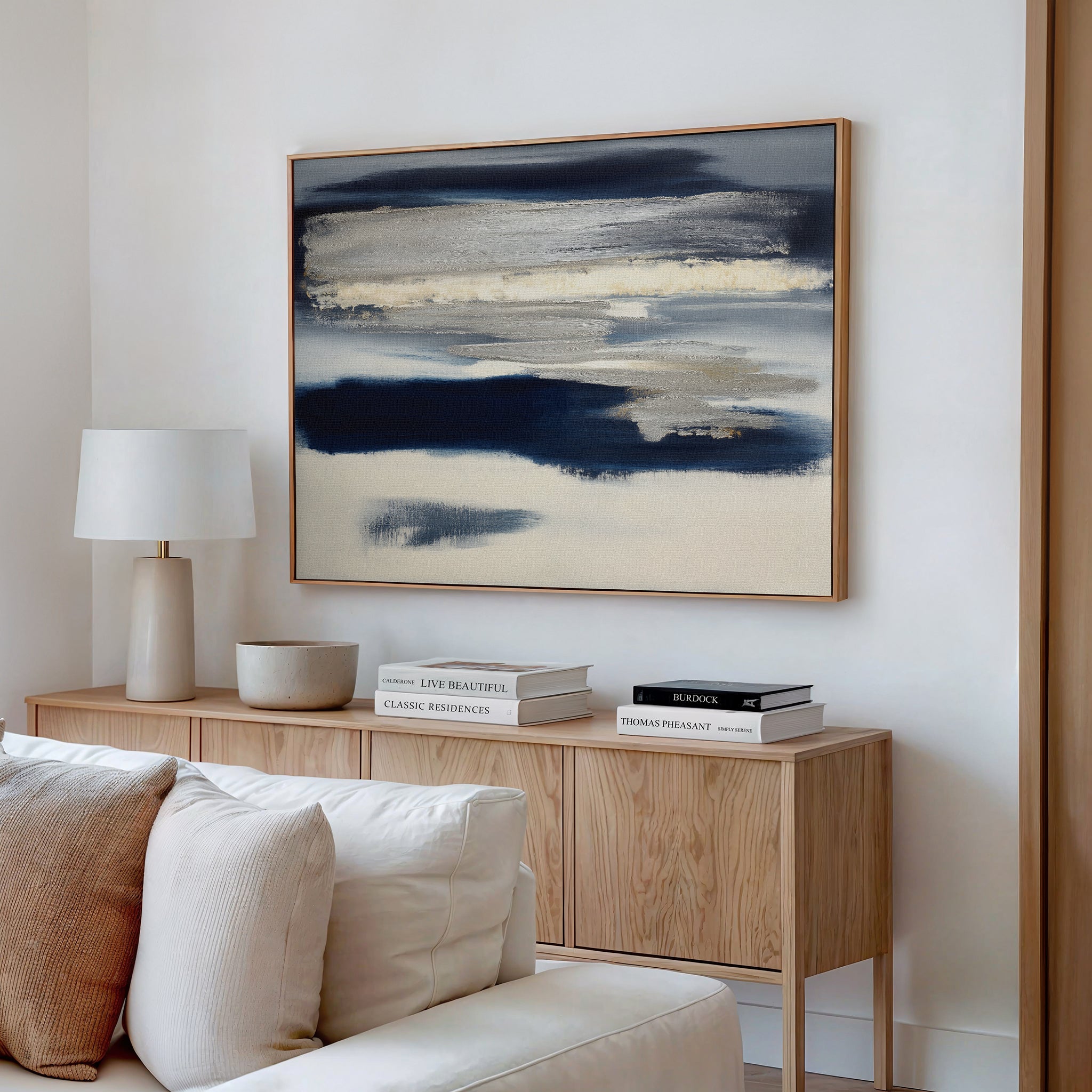 A modern living room features a light wood sideboard, white lamp, stacked books, neutral decor, and the Northern Tides Canvas Art - Horizontal—a striking abstract blue and beige painting—hanging above a cream sofa.