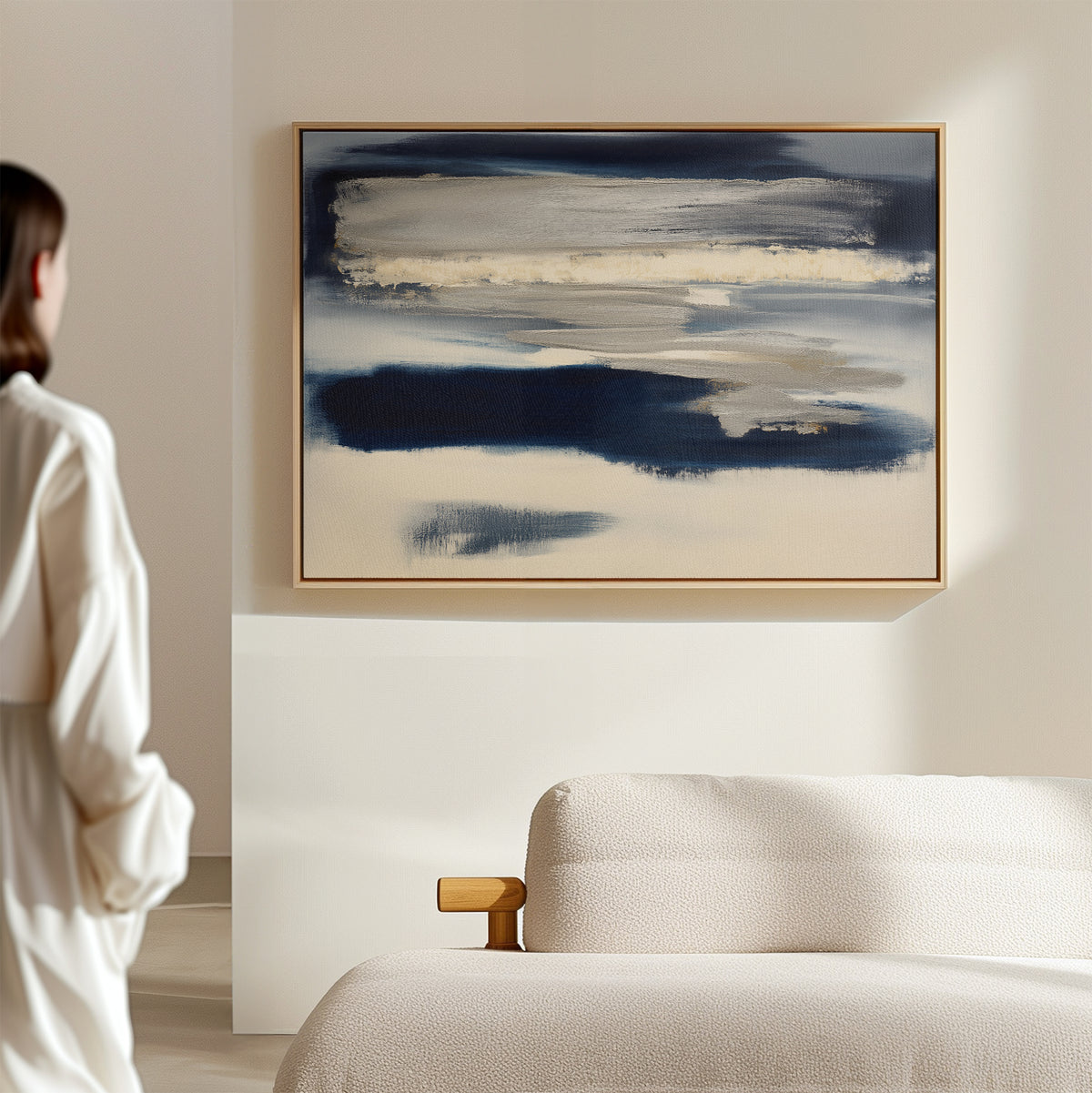 A person stands near a beige sofa, admiring the Northern Tides Canvas Art - Horizontal, which features blue, white, and gray brushstrokes on a light-colored wall in a modern minimalist room.