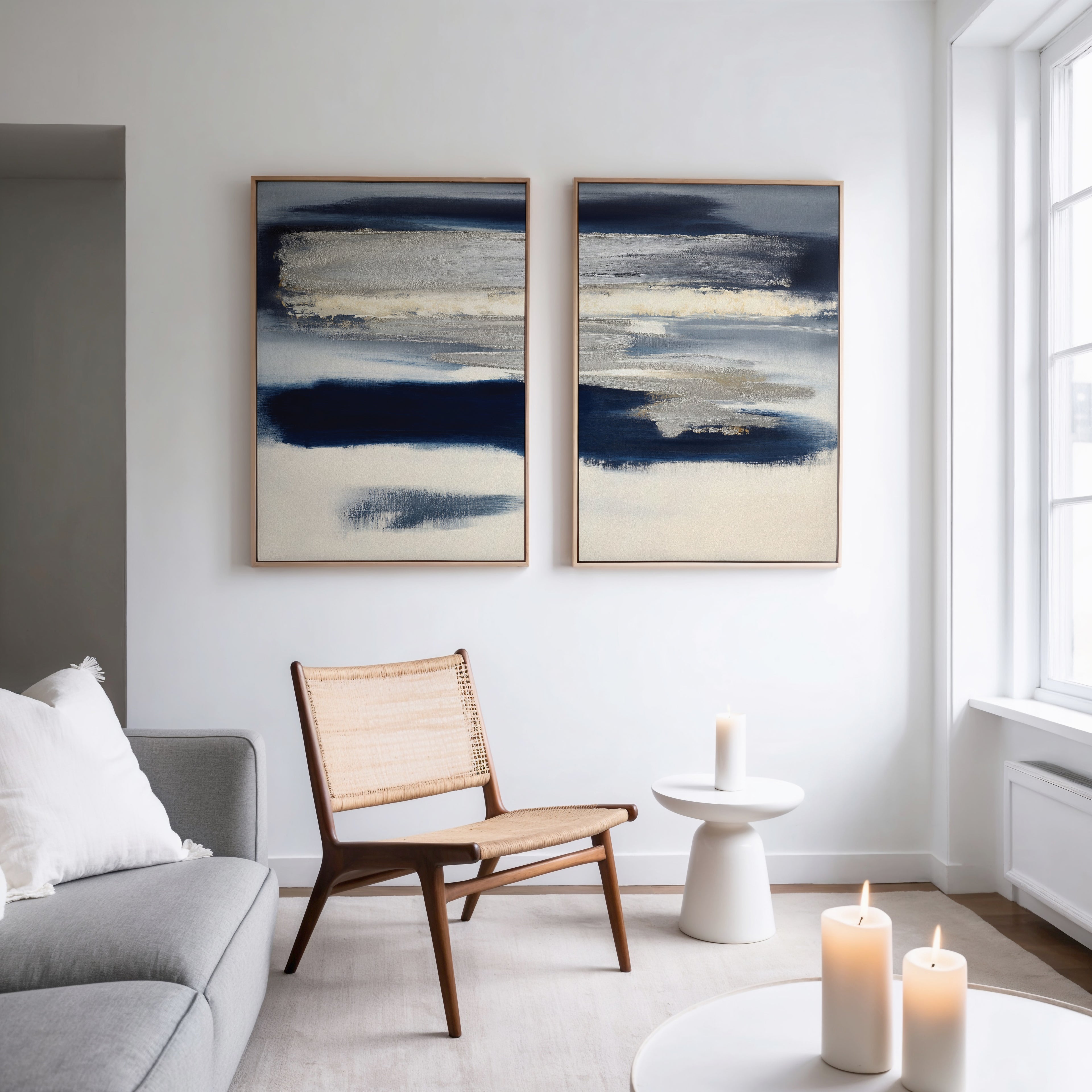 A modern living room with a gray sofa, wooden chair, white table with candle, and Northern Tides - Set Of 2 abstract blue and cream paintings. Minimalist decor and large windows fill the space with natural light.