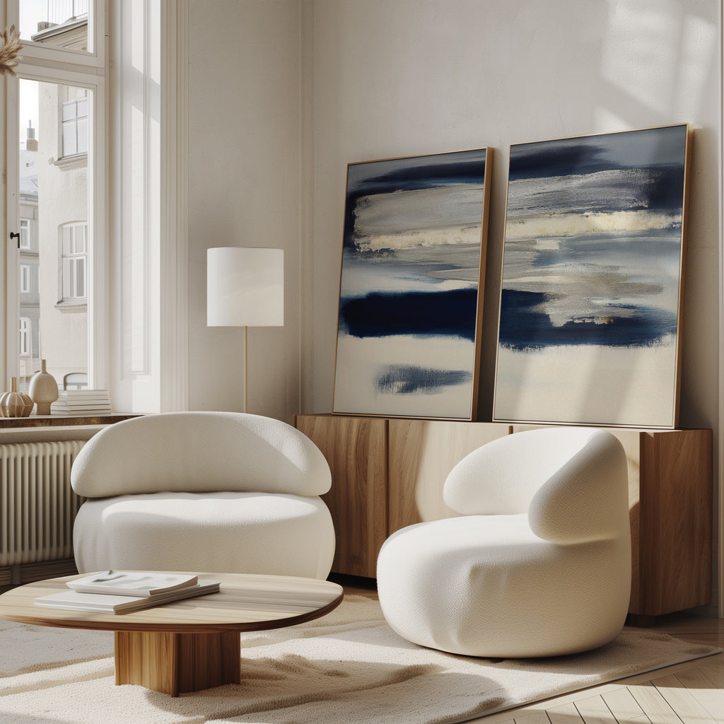 A modern living room features two white, rounded armchairs, a low wooden coffee table, and the Northern Tides - Set Of 2 abstract blue-and-beige artworks on a wooden sideboard. Large windows highlight the minimalist, neutral decor.