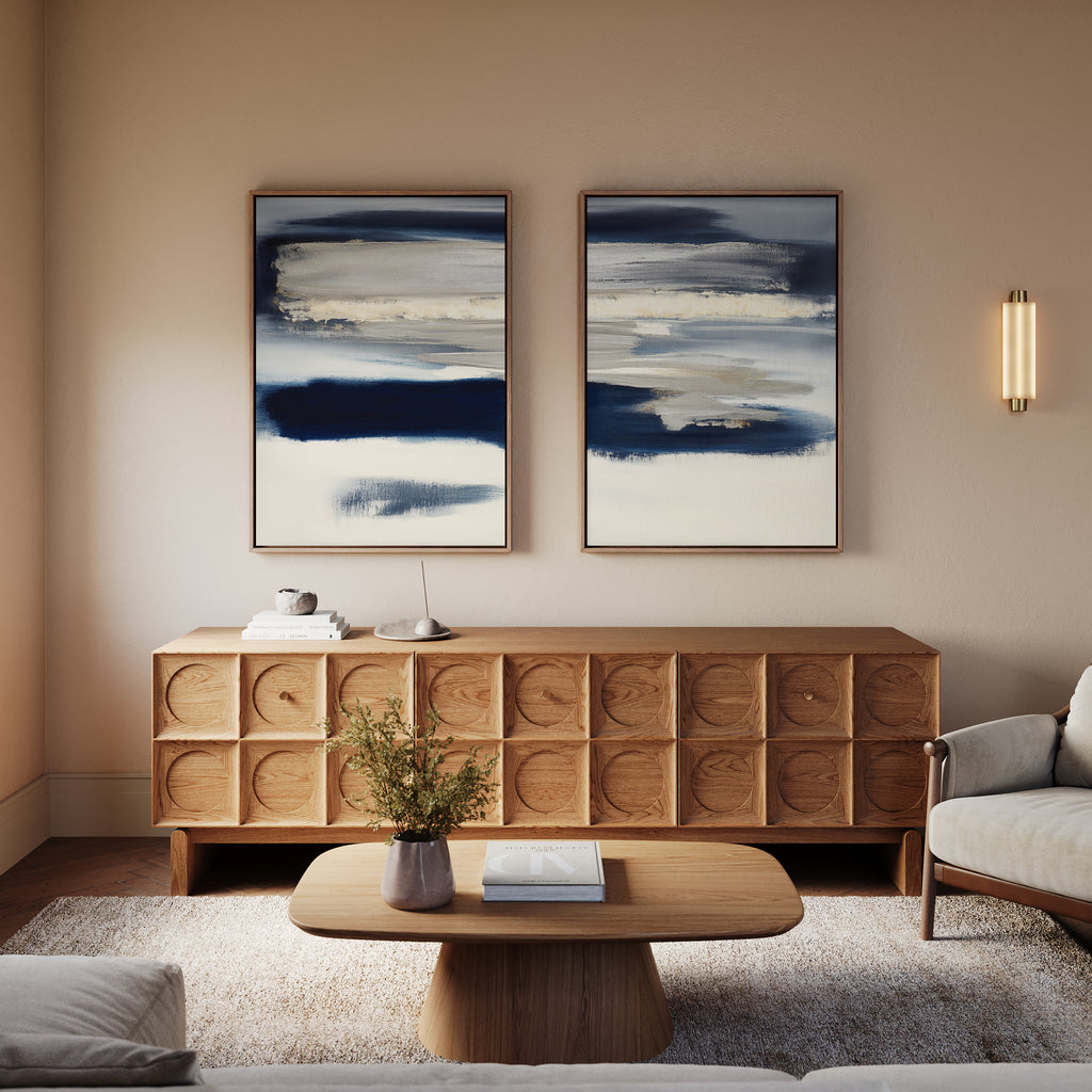 A modern living room featuring the Northern Tides - Set Of 2 abstract blue and white art pieces, a wooden sideboard, an abstract wall print, a round coffee table with a book and vase, light armchairs, and warm ambient lighting.