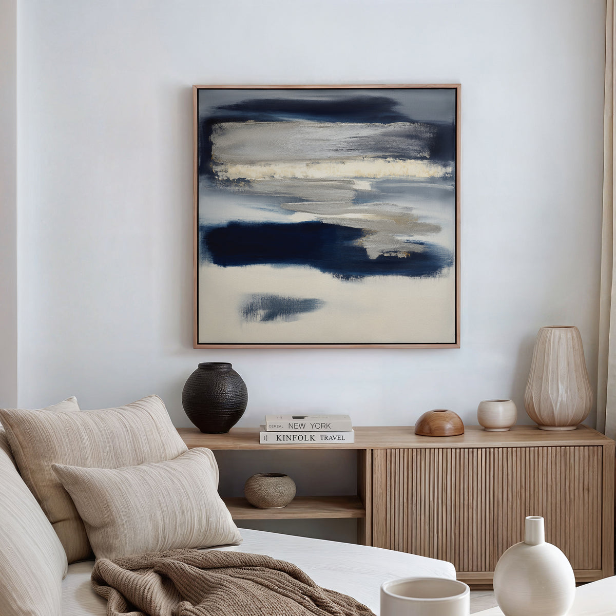 A modern living room featuring a beige sofa, neutral cushions, wooden sideboard, ceramic vases, stacked books, and the Northern Tides Canvas Art - Square as a contemporary indigo abstract print on a white wall.
