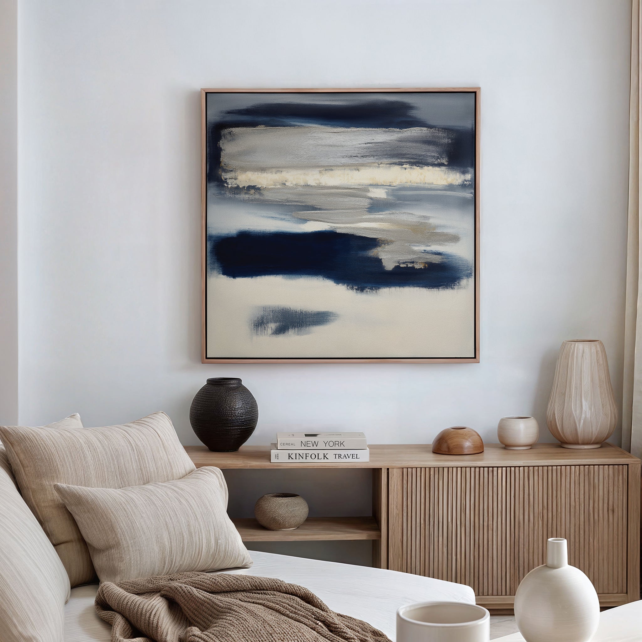 A modern living room featuring a beige sofa, neutral cushions, wooden sideboard, ceramic vases, stacked books, and the Northern Tides Canvas Art - Square as a contemporary indigo abstract print on a white wall.