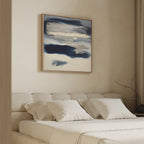 A modern bedroom features a beige upholstered bed with white bedding and pillows. Above the bed is the Northern Tides Canvas Art - Square, showcasing blue and white brushstrokes. A bare branch sits on the bedside table.