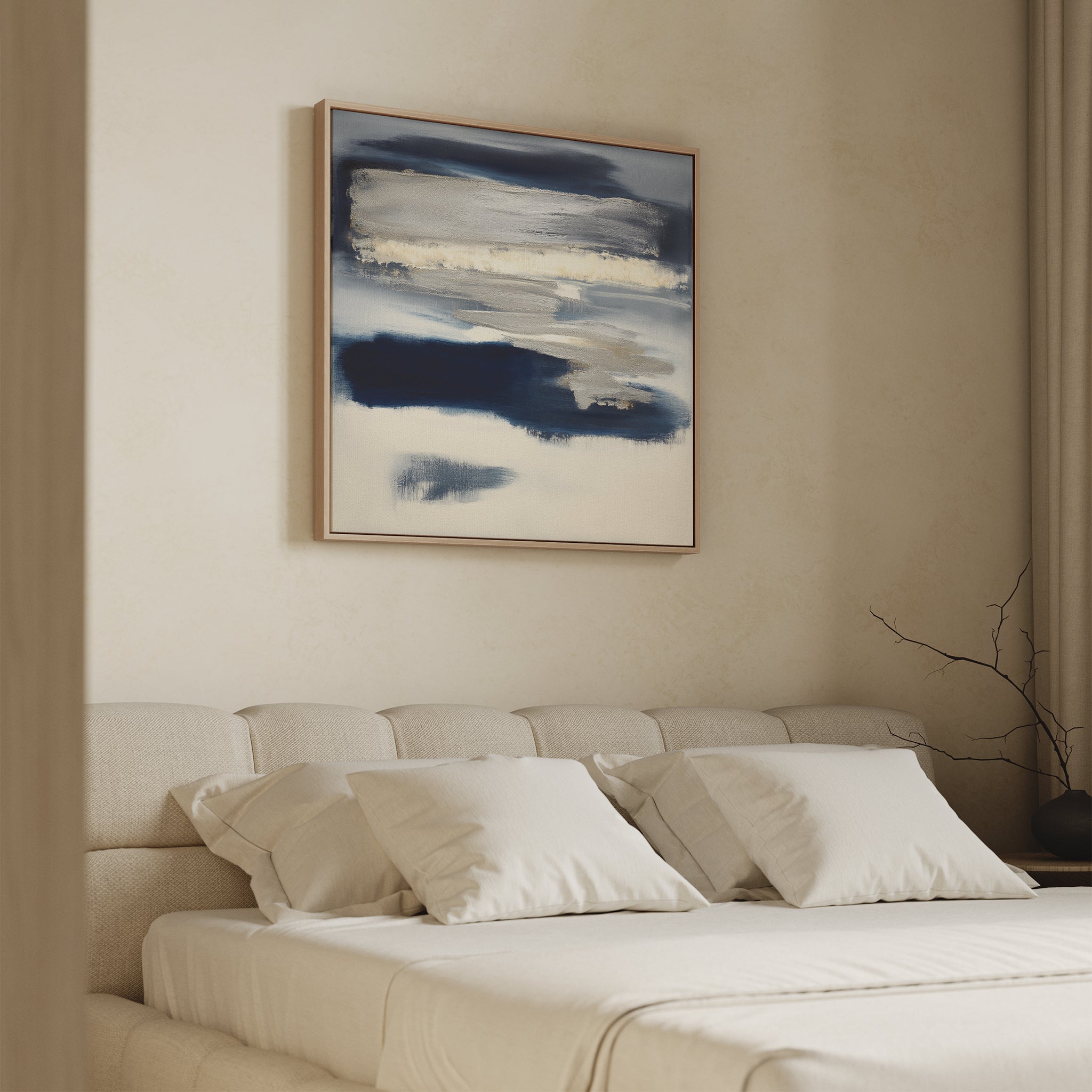 A modern bedroom features a beige upholstered bed with white bedding and pillows. Above the bed is the Northern Tides Canvas Art - Square, showcasing blue and white brushstrokes. A bare branch sits on the bedside table.