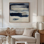 A modern living room with a beige sofa, neutral cushions, side table with ceramic lamp, and a vase of dried flowers. The Northern Tides Canvas Art - Square in indigo blue abstract style hangs above the sofa, adding a contemporary touch.