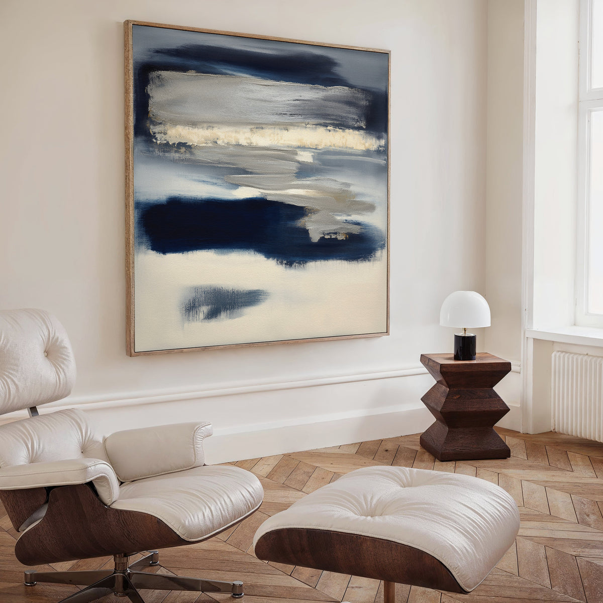 A modern living room featuring a cream lounge chair with ottoman, a small wooden side table with a white lamp, and the Northern Tides Canvas Art - Square showcasing contemporary abstract blue, beige, and white tones as minimalist wall decor.