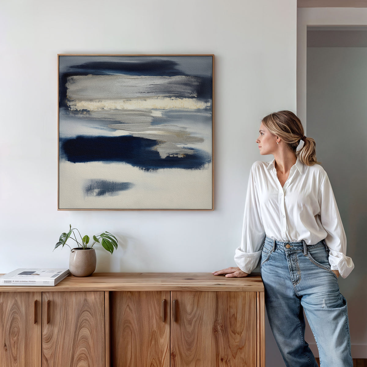A woman in a white blouse and jeans leans against a wooden cabinet, admiring the Northern Tides Canvas Art - Square in indigo blue on the wall. A small plant and book on the cabinet complement the minimalist setting.