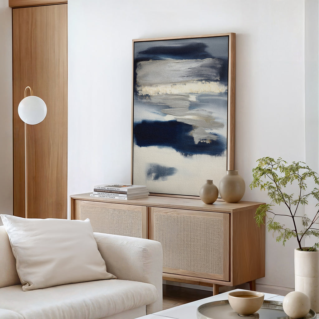 A modern living room with a light wood sideboard, Northern Tides Canvas Art - Vertical in an abstract print, neutral vases, a small green plant, a white sofa with cushions, and a round pendant light against minimalist white walls.
