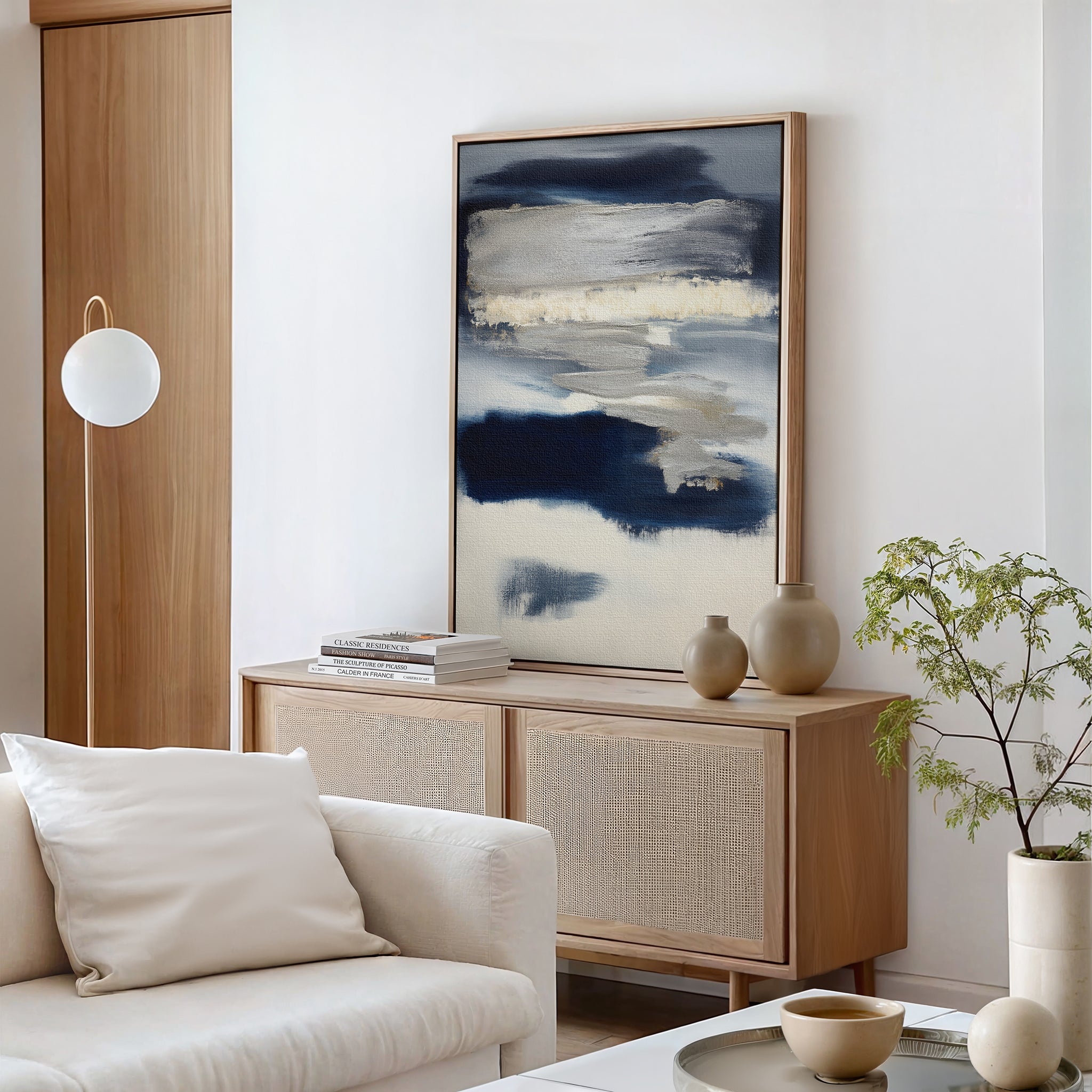 A modern living room with a light wood sideboard, Northern Tides Canvas Art - Vertical in an abstract print, neutral vases, a small green plant, a white sofa with cushions, and a round pendant light against minimalist white walls.