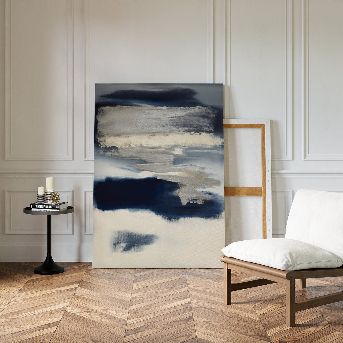 The Northern Tides Canvas Art - Vertical, featuring modern abstract design in navy, cream, and beige, leans against a white paneled wall next to a round black side table with candles and books by a cushioned chair on a herringbone wood floor.