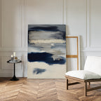The Northern Tides Canvas Art - Vertical, featuring modern abstract design in navy, cream, and beige, leans against a white paneled wall next to a round black side table with candles and books by a cushioned chair on a herringbone wood floor.