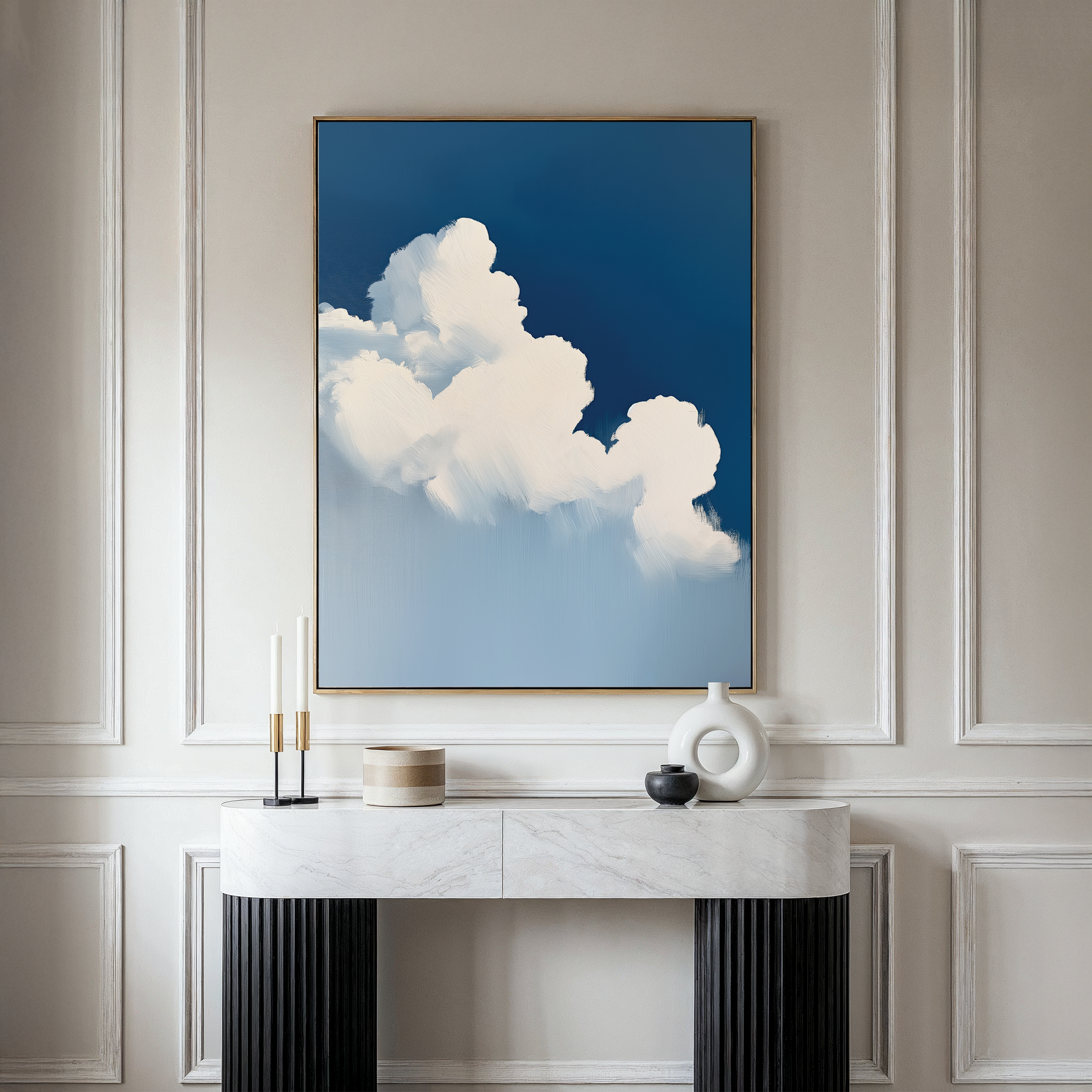 The Cumulus Abstract Canvas Art, a minimalist wall piece depicting white clouds on a blue sky, hangs above a modern marble console table with black legs, complemented by simple vases and candles on a beige wall.