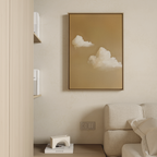A minimalist living space features a beige sofa, shelves with white books, and the framed Whispering Clouds wall art depicting fluffy white clouds on a warm brown background hanging on a light-colored wall.