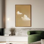 A modern living room showcases Whispering Clouds—a large, minimalist framed painting of white clouds on beige—mounted above a marble ledge with a glass vase of dried flowers, while a green armchair sits in the foreground.