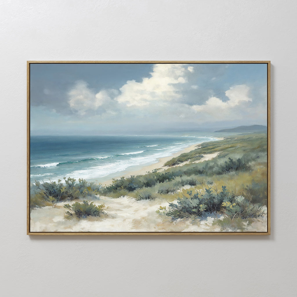 The Ocean Bluff Canvas Art - Horizontal showcases sand dunes, green shrubs, gentle waves, and a blue sky with clouds, framed and displayed on a plain white wall.