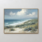 The Ocean Bluff Canvas Art - Horizontal showcases sand dunes, green shrubs, gentle waves, and a blue sky with clouds, framed and displayed on a plain white wall.