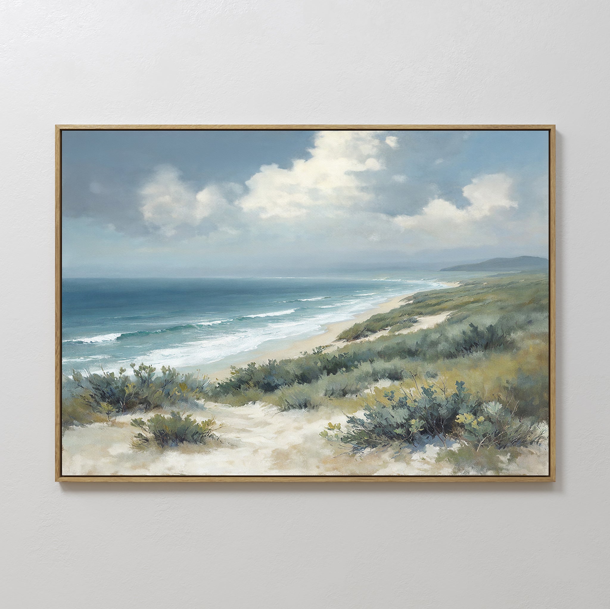 The Ocean Bluff Canvas Art - Horizontal showcases sand dunes, green shrubs, gentle waves, and a blue sky with clouds, framed and displayed on a plain white wall.
