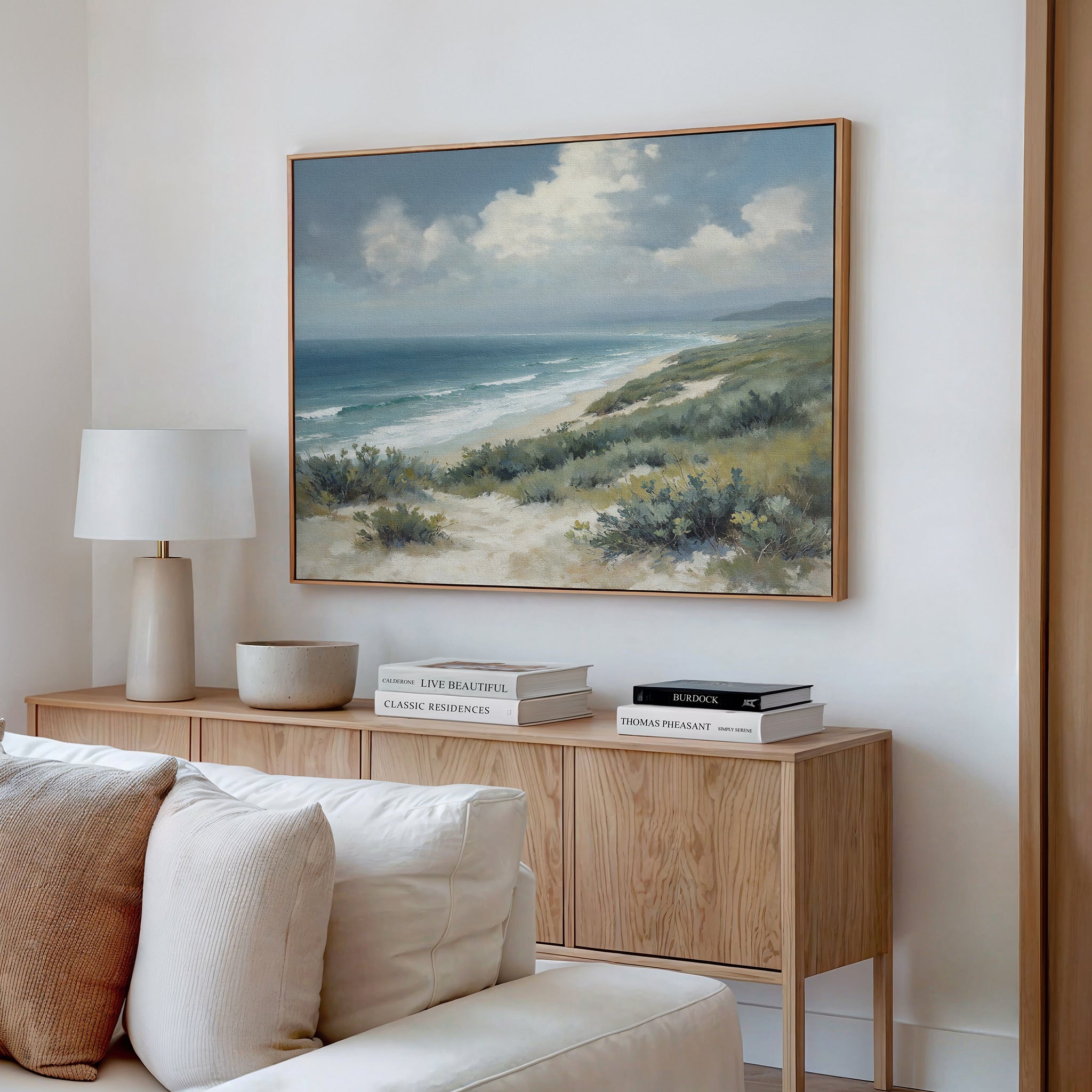 A cozy living room corner features a light wood console table, white lamp, stacked books, and the Ocean Bluff Canvas Art - Horizontal—a large print of sand dunes, bushes, and ocean beneath a partly cloudy sky.