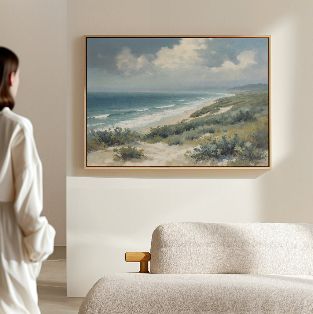 A person in a white robe stands in a softly lit room, admiring the Ocean Bluff Canvas Art - Horizontal featuring sand dunes, bushes, waves, and a cloudy sky, elegantly displayed above a light-colored sofa.