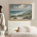 A person in a white robe stands in a softly lit room, admiring the Ocean Bluff Canvas Art - Horizontal featuring sand dunes, bushes, waves, and a cloudy sky, elegantly displayed above a light-colored sofa.