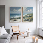 A modern living room features a light gray sofa, wooden chair, and small round table with lit candles. The white wall displays the Ocean Bluff - Set of 2 coastal canvas prints, with more candles on the foreground table.