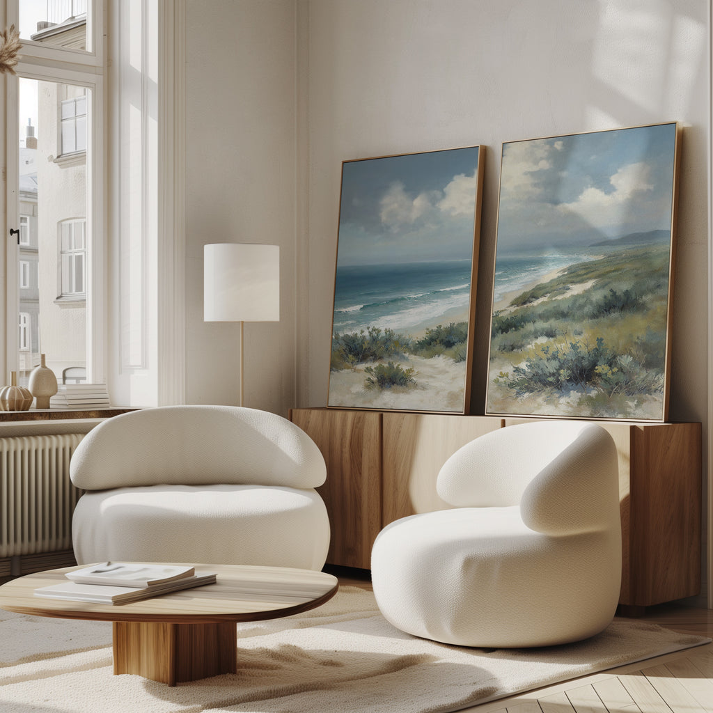 A bright, modern living room with two rounded white chairs, a low wooden table, and the Ocean Bluff - Set of 2 coastal canvas prints leaning against a wooden sideboard under tall windows.