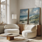 A bright, modern living room with two rounded white chairs, a low wooden table, and the Ocean Bluff - Set of 2 coastal canvas prints leaning against a wooden sideboard under tall windows.