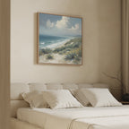 A serene bedroom features a beige upholstered bed with white pillows against a sandy wall. Above the bed hangs the Ocean Bluff Canvas Art - Square, depicting sand dunes, grass, and a blue ocean beneath a partly cloudy sky.
