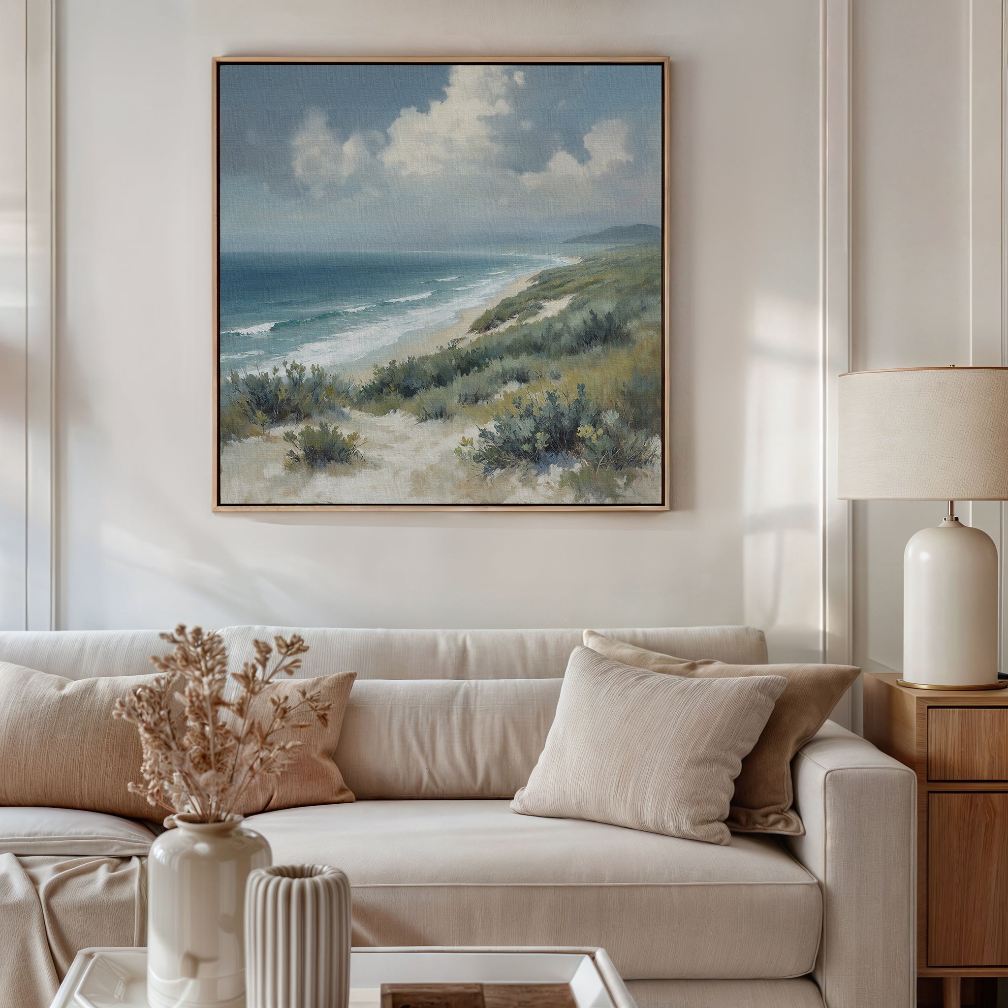 A cozy living room with a cream sofa, beige cushions, a side table with a white lamp, and the Ocean Bluff Canvas Art - Square—a serene seaside print—hanging above the sofa.