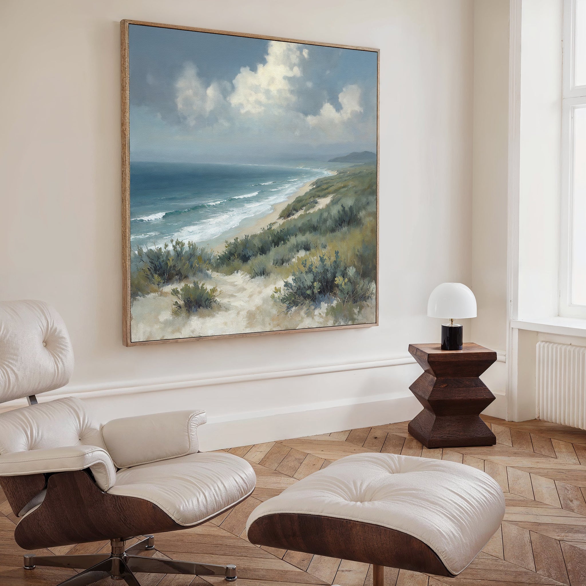 A modern living room with a white lounge chair and ottoman, wooden side table with a white lamp, and the Ocean Bluff Canvas Art - Square hanging above light wood herringbone floors.