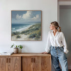 A woman in a white blouse and jeans leans against a wooden cabinet, gazing at the Ocean Bluff Canvas Art - Square, a serene seaside print featuring sand, sea, and clouds, hanging on the white wall above the cabinet.