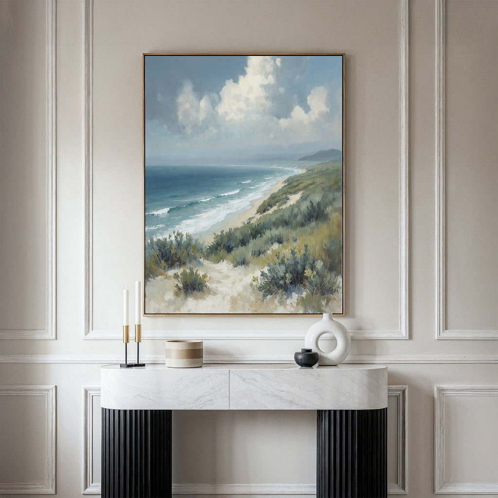 The Ocean Bluff Canvas Art - Vertical hangs on a light-paneled wall above a modern console, showcasing sandy beach dunes, green shrubs, blue ocean waves, and a cloudy sky for a serene coastal vibe.