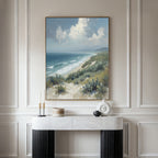 The Ocean Bluff Canvas Art - Vertical hangs on a light-paneled wall above a modern console, showcasing sandy beach dunes, green shrubs, blue ocean waves, and a cloudy sky for a serene coastal vibe.