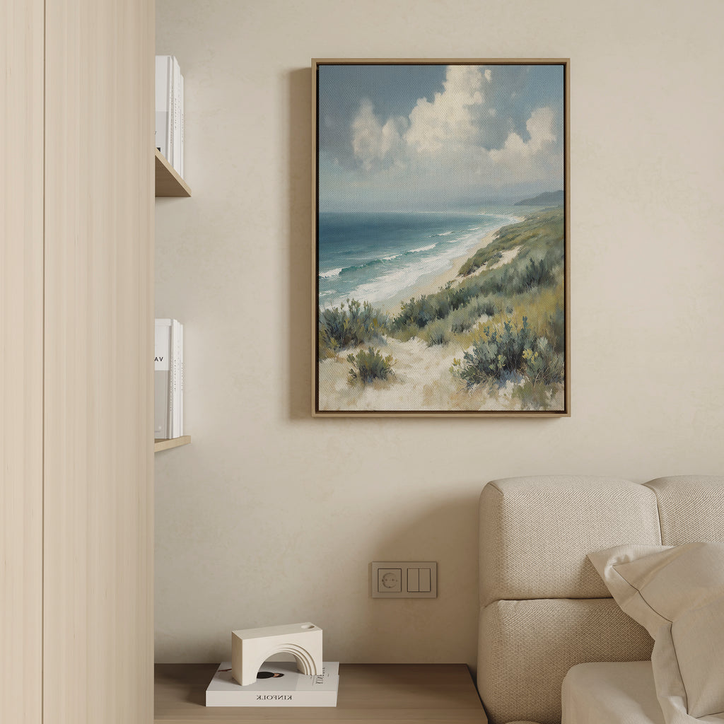 The Ocean Bluff Canvas Art - Vertical hangs above a light-colored sofa, showcasing sand, green shrubs, waves, and a cloudy sky. Shelves with books and decor nearby complete this tranquil coastal living room scene.