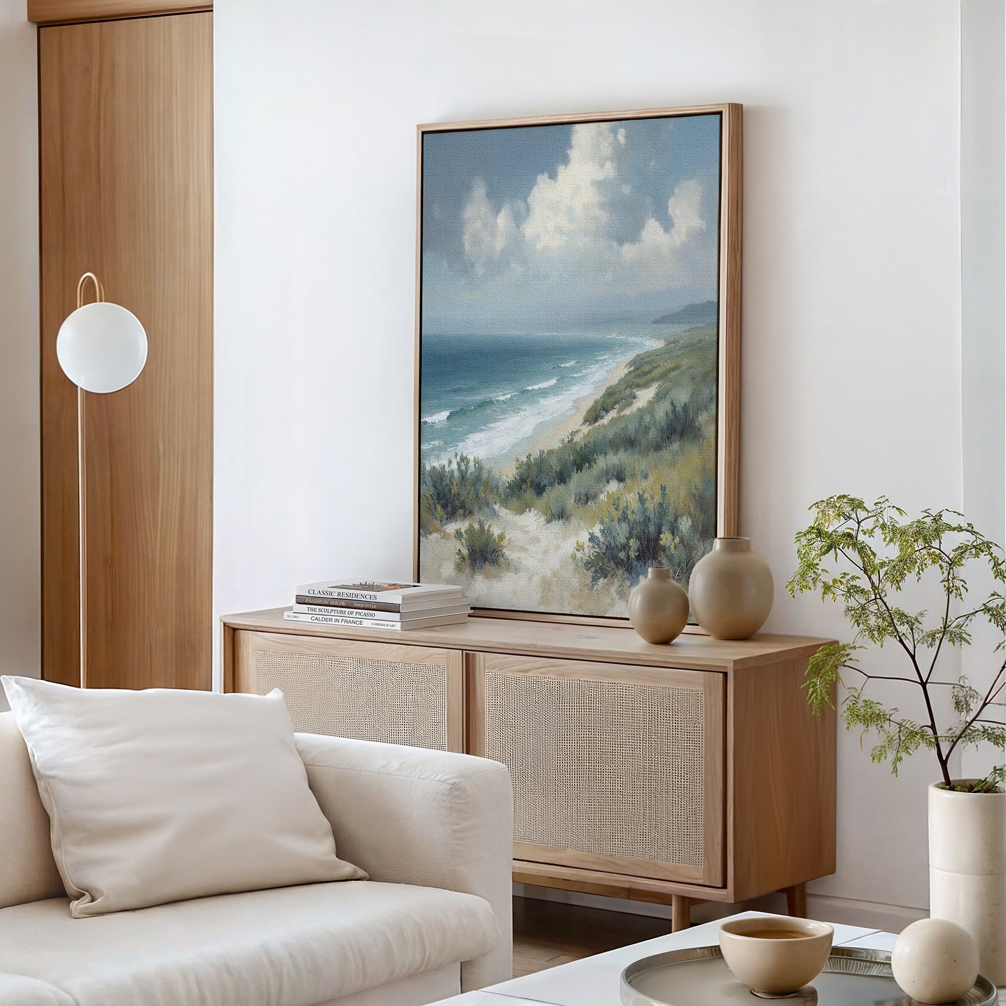 A minimalist living room featuring the Ocean Bluff Canvas Art - Vertical above a light wood sideboard, stacked books, a round white lamp, a beige couch, a small plant, and neutral ceramic decor on the white coffee table.