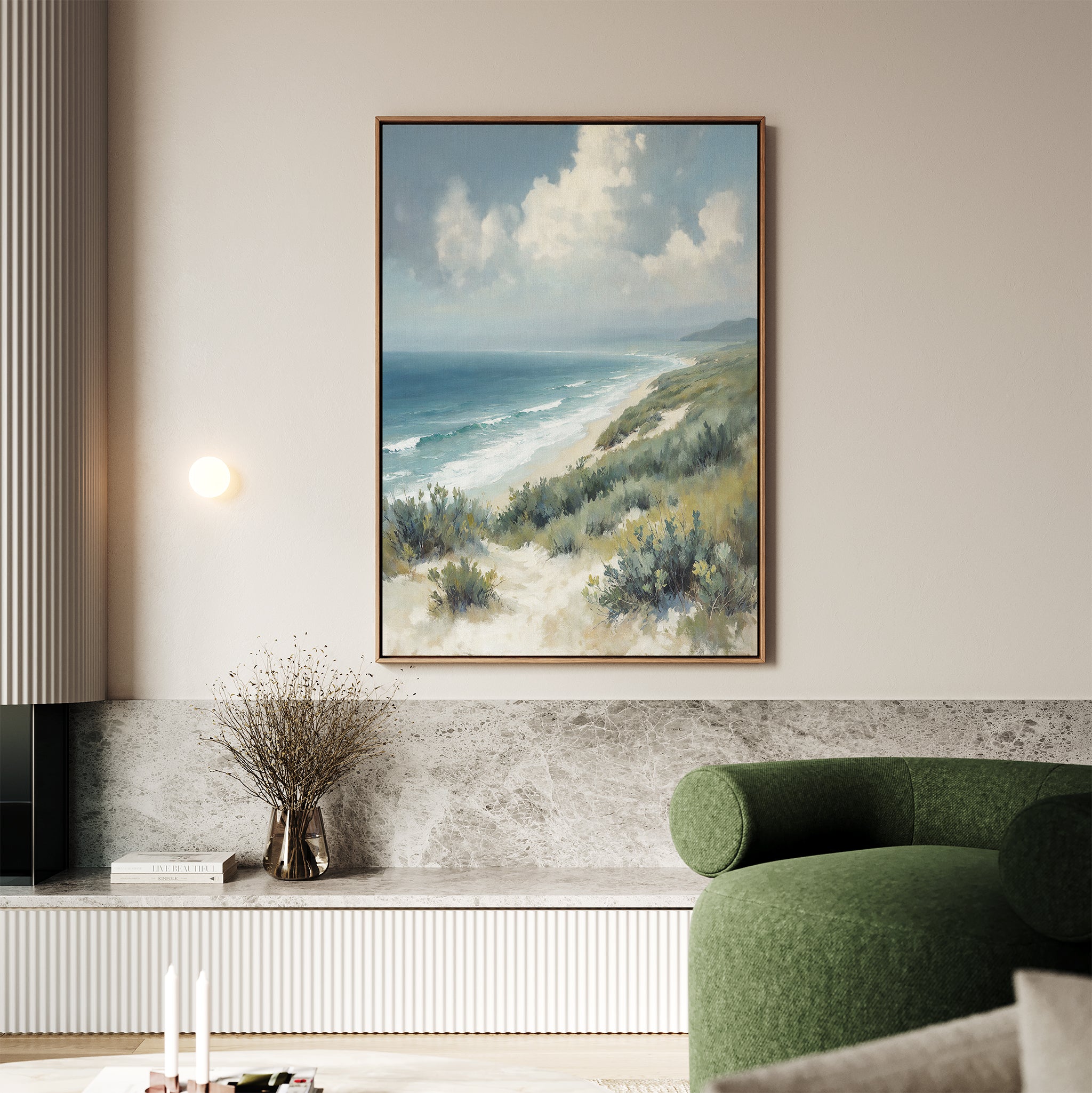 A living room with a light green curved sofa, marble-topped bench, dried flower vase, and the Ocean Bluff Canvas Art - Vertical, featuring sand dunes and ocean beneath a cloudy sky, adds serene seaside style to the space.