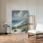 The Ocean Bluff Canvas Art - Vertical leans against a white paneled wall, beside a white cushioned chair and a black table with books and candles on parquet wood flooring, creating a serene seaside atmosphere.