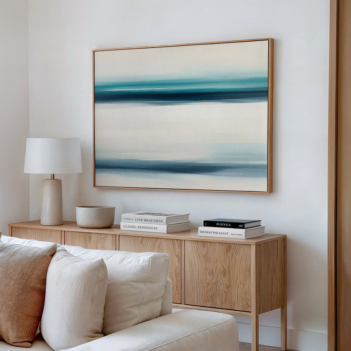A minimalist living room features a light wood sideboard, white lamp, decorative bowl, and stacked books. Above the cream sofa, the Ocean Motion Canvas Art - Horizontal brings a serene coastal vibe with its blue and beige abstract lines.