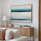 A minimalist living room features a light wood sideboard, white lamp, decorative bowl, and stacked books. Above the cream sofa, the Ocean Motion Canvas Art - Horizontal brings a serene coastal vibe with its blue and beige abstract lines.