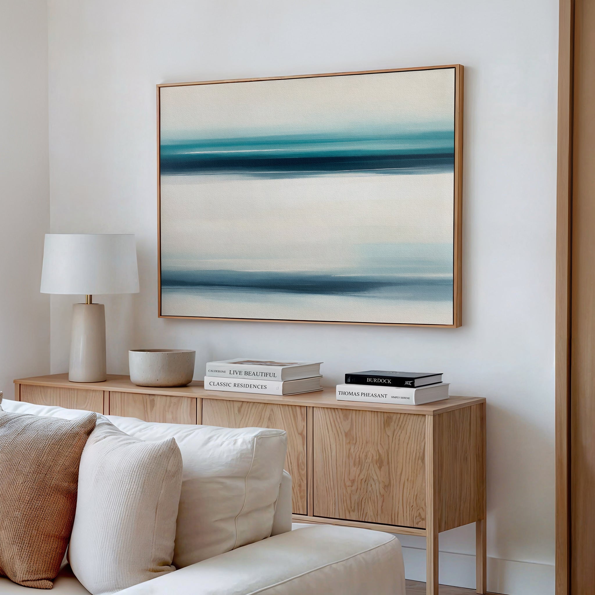 A minimalist living room features a light wood sideboard, white lamp, decorative bowl, and stacked books. Above the cream sofa, the Ocean Motion Canvas Art - Horizontal brings a serene coastal vibe with its blue and beige abstract lines.