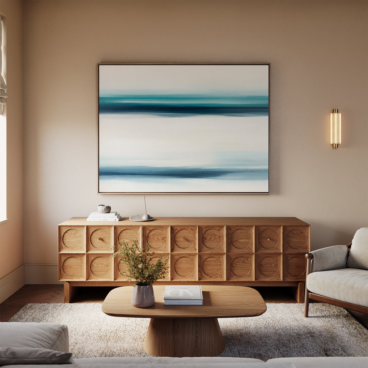 A modern living room features a wooden sideboard topped by the Ocean Motion Canvas Art - Horizontal, a small round coffee table with flowers and a book, a light rug, and a beige sofa. Warm lighting enhances the serene, sophisticated atmosphere.