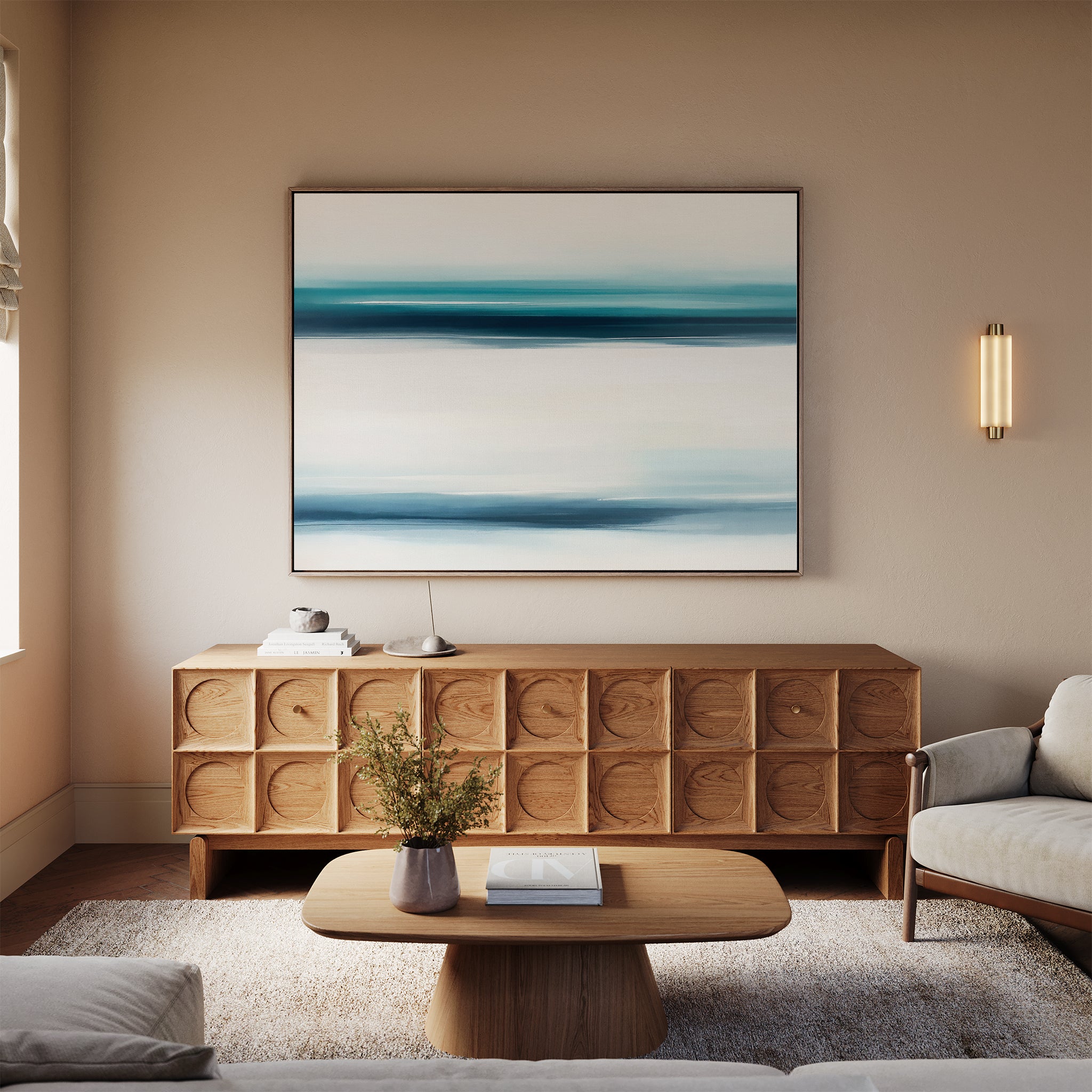 A modern living room features a wooden sideboard topped by the Ocean Motion Canvas Art - Horizontal, a small round coffee table with flowers and a book, a light rug, and a beige sofa. Warm lighting enhances the serene, sophisticated atmosphere.