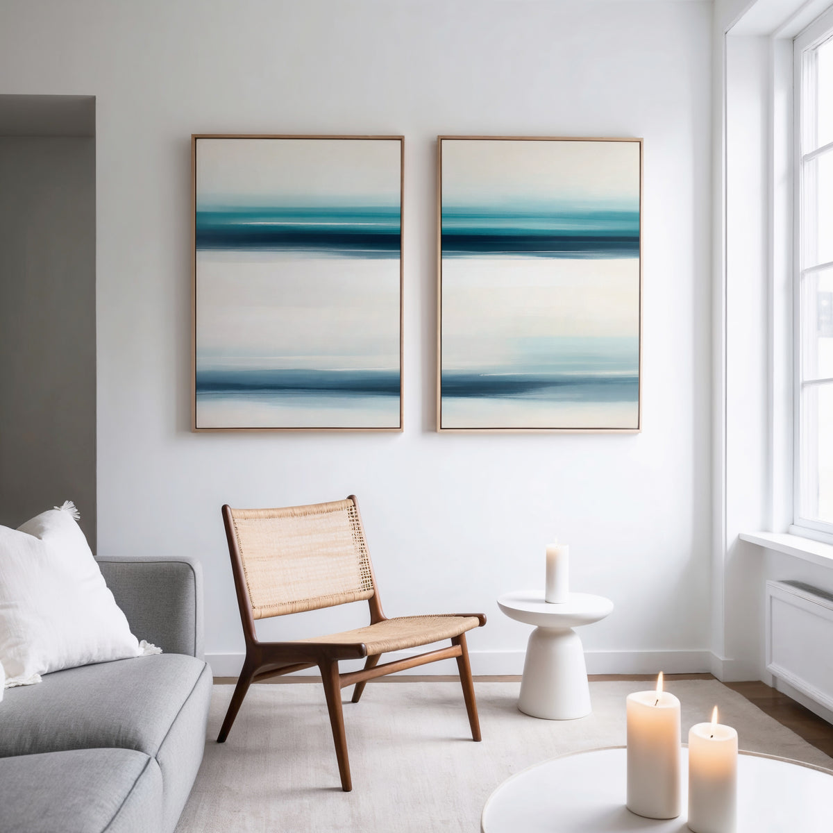 A modern living room with a gray sofa, woven chair, and small white side table holding a candle exudes serene sophistication. The Ocean Motion - Set Of 2 art adds coastal flair while large windows fill the space with natural light.